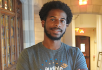 Audible Software Engineer Justin Cockburn stands in our Innovation Cathedral in Newark, the former church turned office's stained glass behind him. He stands center frame, looking straight to camera, smiling, wearing a gray t-shirt with the Audible logo on it.