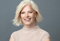 A headshot of Susan Jurevics, Audible's EVP, Head of International. Her head and shoulders are shown in close up, against a light gray background. She is looking directly to camera, smiling and wearing a light rose and white sweater.