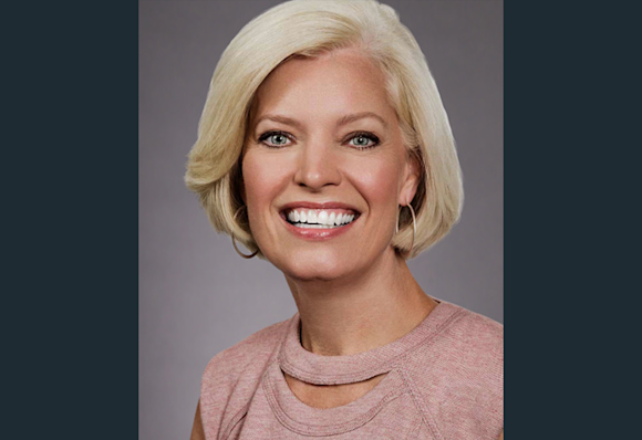 A headshot of Susan Jurevics, Audible's EVP, Head of International. Her head and shoulders are shown in close up, against a light gray background. She is looking directly to camera, smiling and wearing a light rose and white sweater.