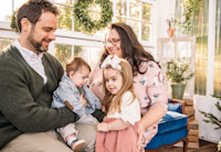 Audible employees Jessica and Joe Reese sit on a blue velvet couch in front of a picture window decorated with a holiday wreath holding their two small children in their laps. They are looking down at their children and smiling.