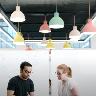 An Audible employee and a tech intern sit at a table working on a project together in our Innovation Cathedral in downtown Newark where a lot of our tech teams work. There are two whiteboards set up behind them. Large colorful pedant lights hang from the ceiling above them.