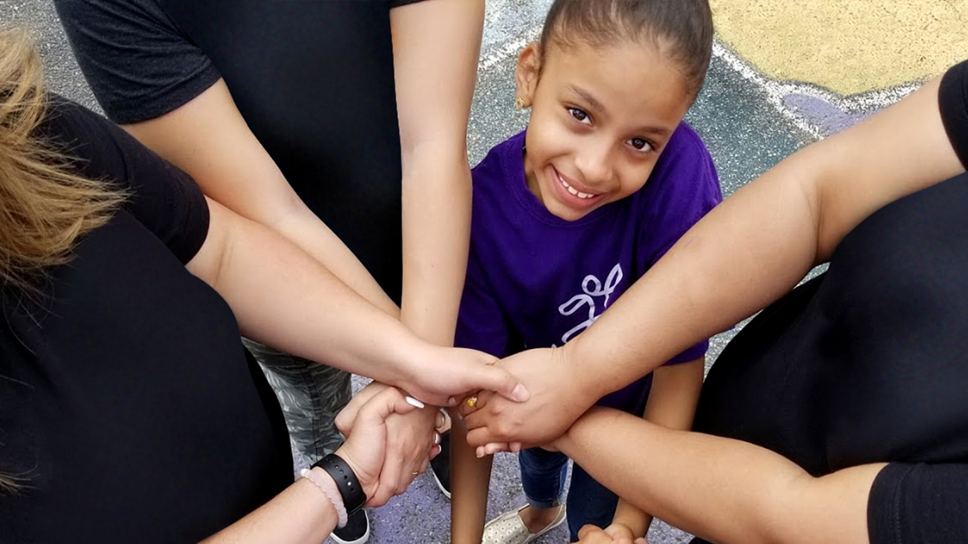 A group of Audible employees stand in a circle with a little girl during a volunteer event, their arms crossed over each other to form a ring of held hands at the circle's center. The photo is taken from the top down so the adult volunteers are seen as black t-shirts and hands and partial faces while the girl is clear at the top of frame and looking up and smiling. She is, frankly, adorable.