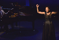 Alexandra Silber, wearing a black dress, sings at a microphone center stage. A man is playing a baby grand piano behind her.