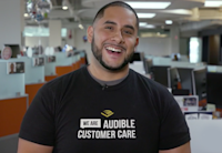 A smiling Raphael stands in Audible Customer Care center wearing a customer care shirt.