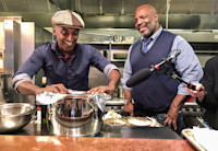 Chef Marcus Samuelsson at the stove of one of his restaurants with a guest standing by him smiling.