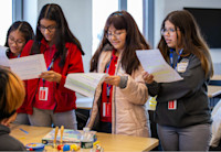 Four young middle-school-aged girls are holding a worksheet and reviewing the scribbled writing on the page. They're in what appears to be a small conference room located in Audible's 1 Washington offices.