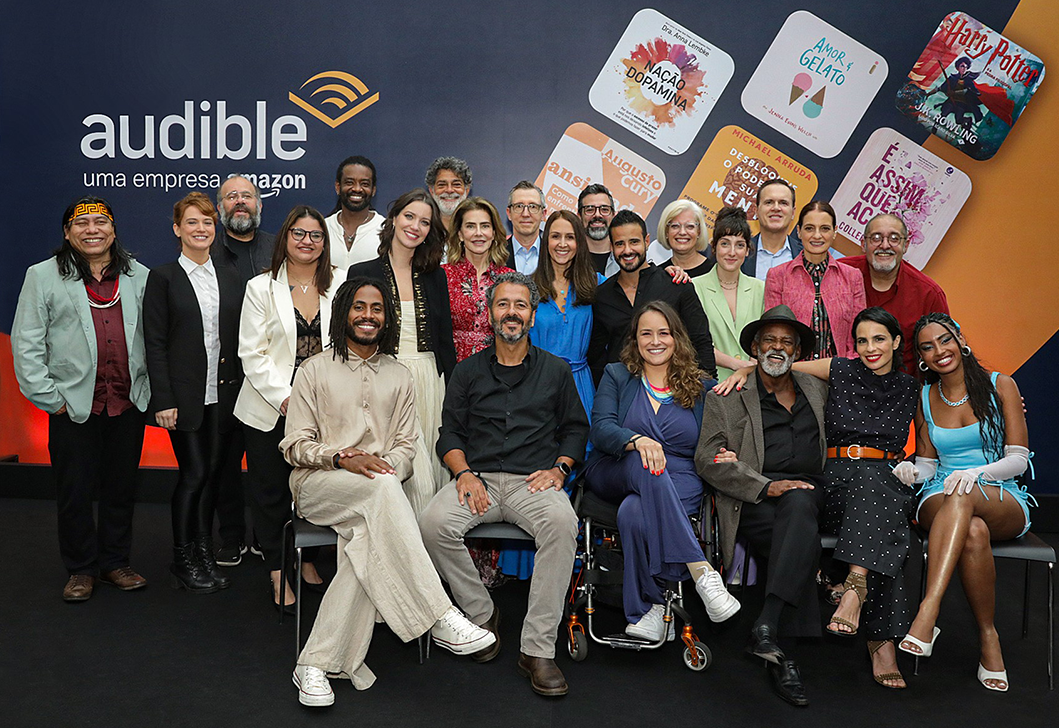 A group of actors and Audible employees gathers together in front of a backdrop with audiobook cover art and the Audible Brazil logo.
