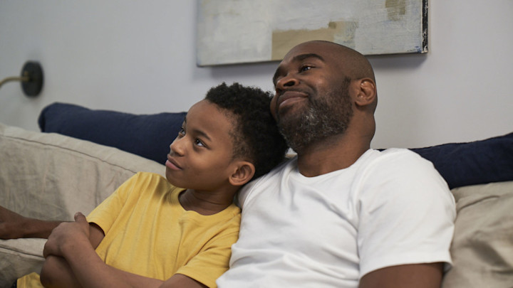 A man sits on a couch with his arm around his son. They appear to be relaxing and listening to something together.