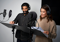 A photo of Cobie Smulders and Raymond Ablack in the studio. They're wearing headphones and reading lines from a script.