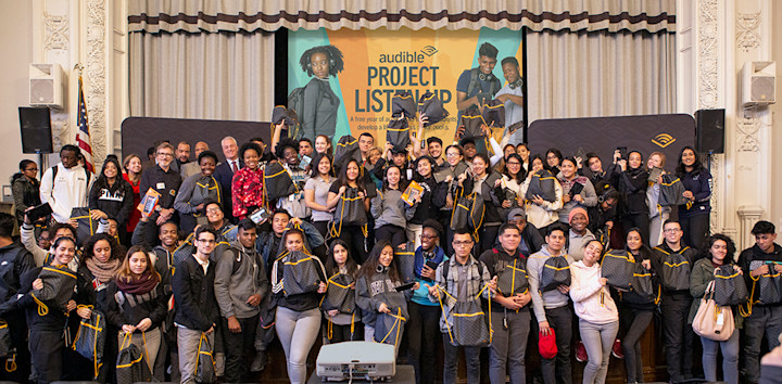 A large group of students and Audible employees stand on front of and on top of a stage excitedly holding bags filled with tablets and earphones to celebrate the launch of Project Listen Up.