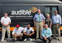 Six people standing or squat in front of one of the vans that is part of their shuttle company in Newark, NJ. The van has Audible's name and logo on it and the tagline, "proudly headquartered in Newark."