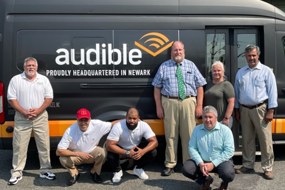 Six people standing or squat in front of one of the vans that is part of their shuttle company in Newark, NJ. The van has Audible's name and logo on it and the tagline, "proudly headquartered in Newark."