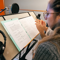 A printed script sits on a stand, a mic behind it and an actor before it, her face in profile.