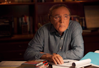 The best-selling author James Patterson sits behind his desk staring off camera. His arms are crossed and propped up on the desk. A pad of paper sits on his desk, a pen lying across it by his hand.