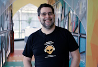 Oren Levin stands straight at the camera smiling and wearing a black shirt that features an alien wearing headphones and the words "Audible Engineering" above it. Behind him is a colorful backdrop of Audible's lobby at its Newark headquarters.