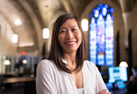 Audible’s CFO and Growth Officer Cynthia Chu is sitting in Audible's Innovation Cathedral. She is wearing a white sweater and looking at the camera smiling. Behind her you can see stained glass windows.