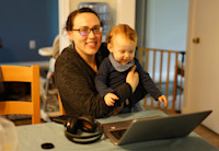 A woman sits at her kitchen table, her son, a toddler, held in her lap, a laptop open on the table in front of her.