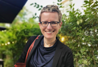 Audible employee Sibylle Schwarz stands in the outdoor patio of a restaurant with greenery behind her, black table umbrellas overhead, and lights strung across the background. She is looking directly to the camera and smiling. She has short cropped blond hair and is wearing dark framed glasses, a navy shirt and a black blazer. She has a red purse slung over her shoulder.