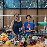 Two people stand behind a table piled with goods for sale.