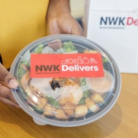 Person holding to-go container filled with salad, with a red NWK Delivers logo.