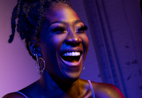Amber Iman smiles and is glowing with a purple hue coming from a spotlight.