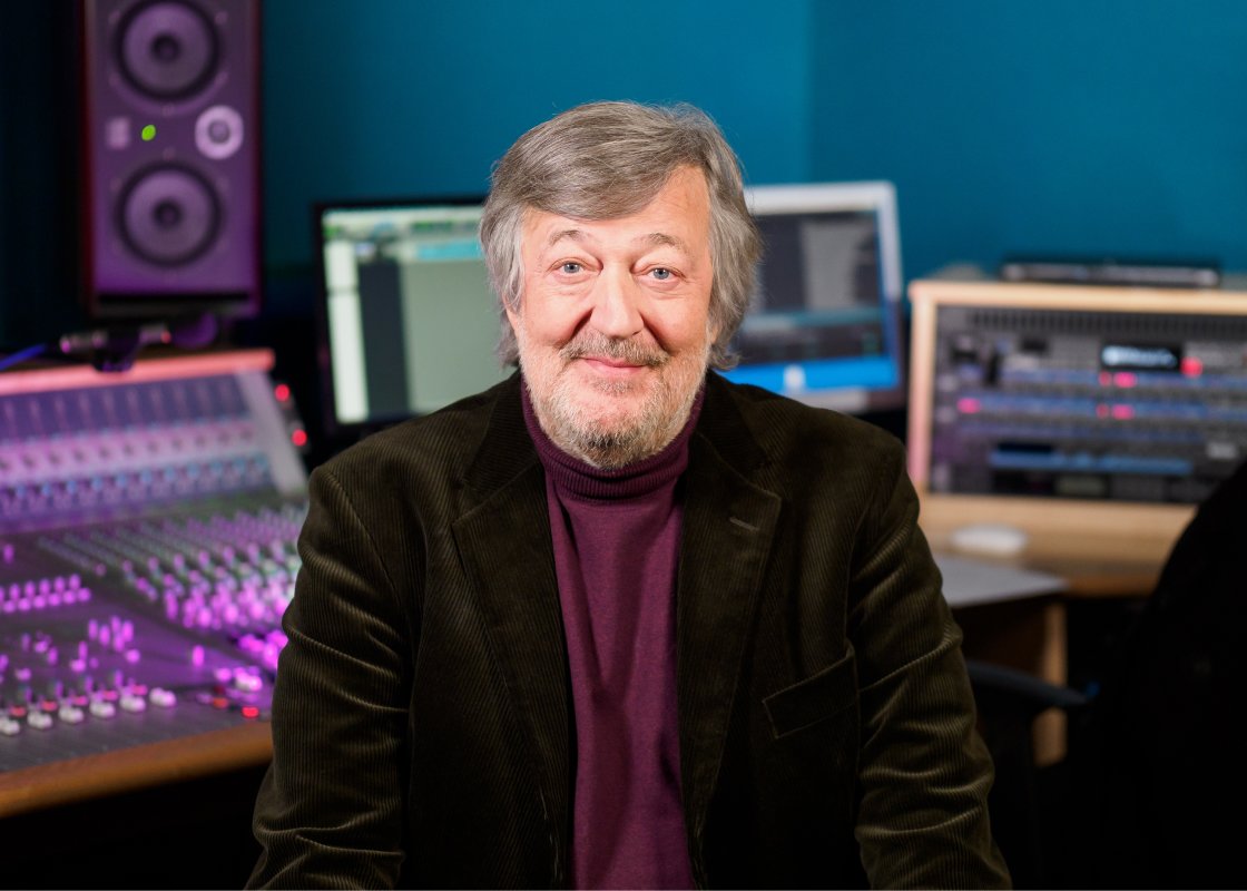 Actor, writer and Audible narrator Stephen Fry sits in an Audible recording studio. A mixing board, computer monitors and a speaker are behind him.