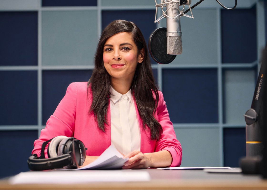 Collien Ulmen-Fernandes sits in a recording studio in front of a microphone, smiling to camera.