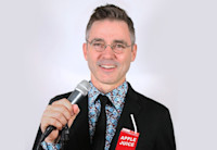 Comedian Billy Kelly, wearing wire-framed glasses, a blue and purple patterned shirt, black tie and suit jacket, stands holding a microphone in his right hand and a apple juice box with straw in his left hand.