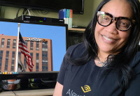 Denise Davis wearing black rimmed glasses and an Audible t-shirt sits in front of a computer monitor showing a photo of Audible's Newark headquarters. 