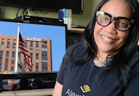 Denise Davis wearing black rimmed glasses and an Audible t-shirt sits in front of a computer monitor showing a photo of Audible's Newark headquarters. 