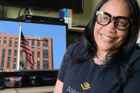 Denise Davis wearing black rimmed glasses and an Audible t-shirt sits in front of a computer monitor showing a photo of Audible's Newark headquarters. 
