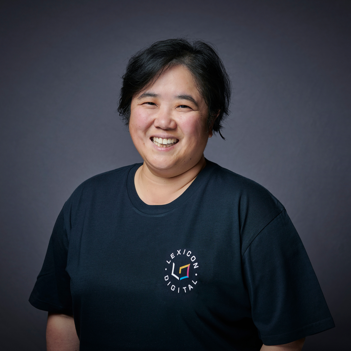 A photograph of Catherine Thum, smiling direct to camera. Behind her is a black wall. Catherine is wearing a black t-shirt, with a logo. The logo says Lexicon Digital.