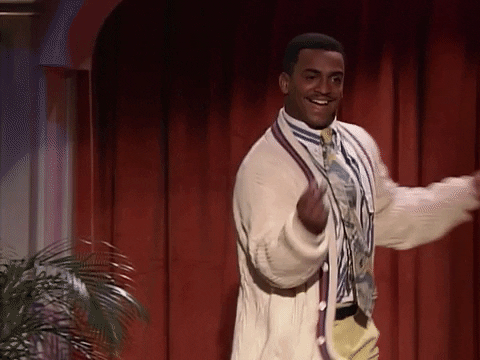 A GIF image of Alphonso Ribeiro dancing. He is an African American actor who starred in The Fresh Prince of Bel-Air. He is wearing cream trousers with a brown belt, a white shirt with black stripes, a cream cardigan with red borders, and a long tie with yellow graphics. He is dancing side to side with his arms swinging up and down while smiling. He is dancing on a stage, against a backdrop of red curtains. There is a plant to his left, just out of frame. A female figure with short black hair and dangling silver earrings appears briefly in the middle of the GIF and seems to be dancing too. 