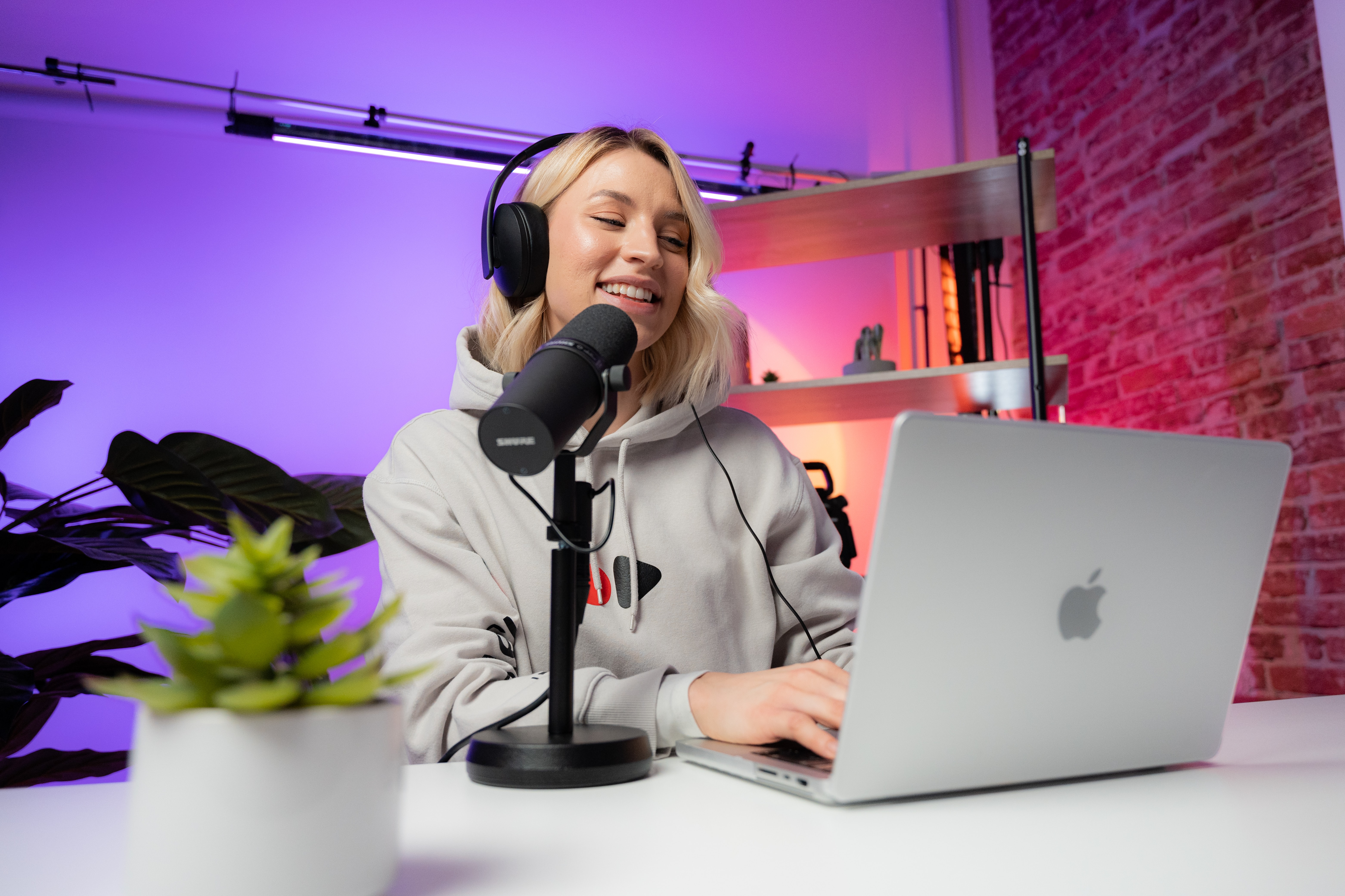 A photograph of a fair-skinned, woman with light blonde hair sitting at a wooden desk. She is smiling and working on a silver Mac laptop. She wears a silver hoodie. She has on a set of black headphones and appears to be speaking into a black microphone. Beside the laptop is a small green plant in a white pot. Behind her is a wall lit by purple light and just out of frame is a larger plant in one corner. In the other corner is a wooden shelf against a red brick wall.