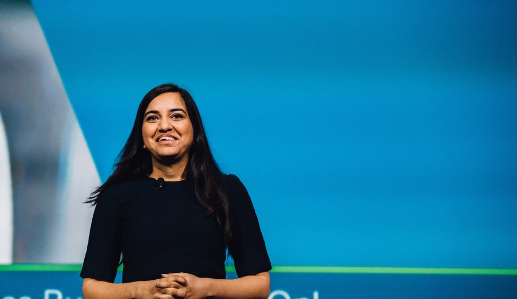 A photograph of Kriti Sharma, a brown skinned woman with long black hair. She is wearing a black top with a microphone attached to the collar, and appears to be delivering a keynote speech. She is well lit and smiling in front of a blue backdrop, which appears to be a slideshow presentation, given the obscured text just out of frame. She is looking up and out at what we assume is an audience, with her hands knitted together in front of her.