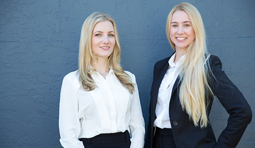A photograph of Jessica and Karina, the founders of DataMinds. Both women are standing next to each other against the backdrop of a dark grey wall. Both are smiling, looking direct to camera. Jessica is standing to the left. She has mid-length blond hair and is wearing a white shirt with black trousers. Karina has long blond hair, and is wearing a white shirt underneath a black blazer with black trousers. She has her hand in her pocket.