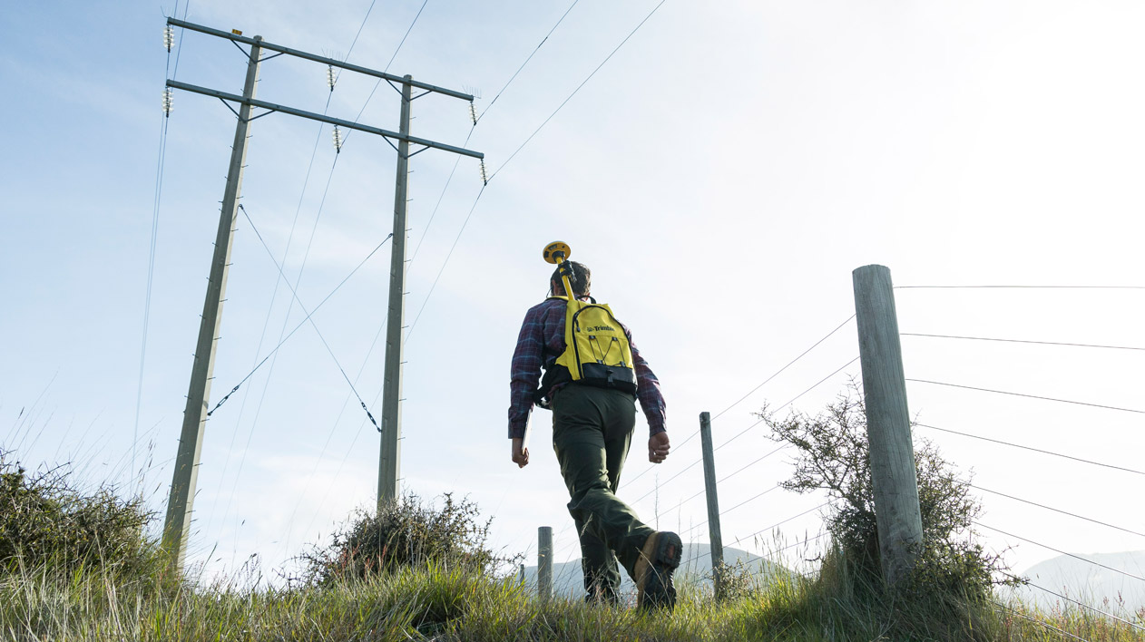 A person with a bright yellow. Trimble backpack and surveying equipment walks up a grassy hill towards large power lines and utility poles against a clear sky.