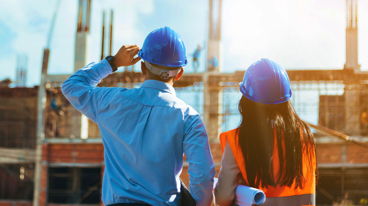 Two individuals in blue hard hats and safety vests stand with their backs to the viewer, observing a construction site with partially built structures under a bright sky.