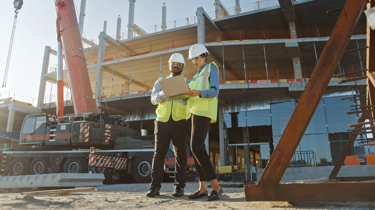 Two construction managers in hard hets & reflective vests look at computer on job site in front of building under construction.