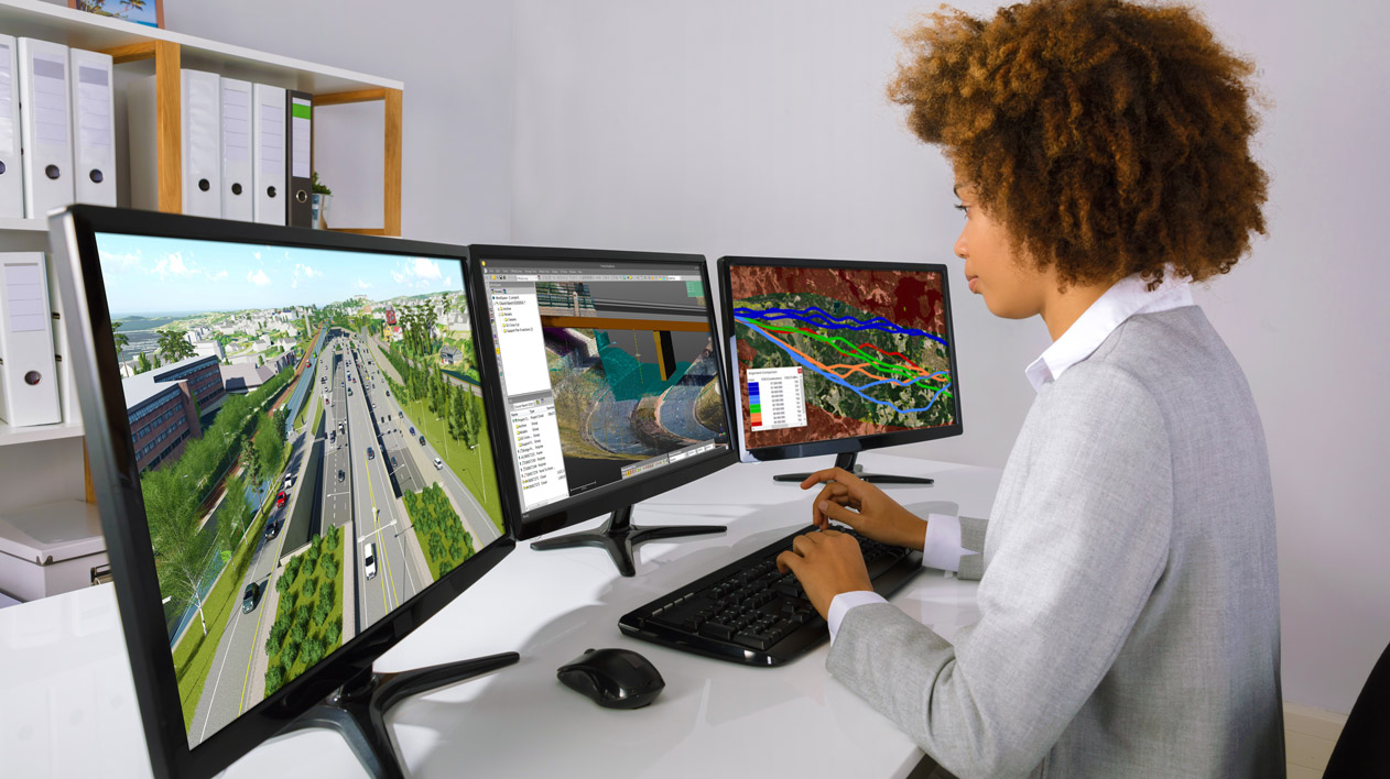 A woman with curly hair sits at a desk, looking at three computer monitors displaying 3D models and data visualizations.