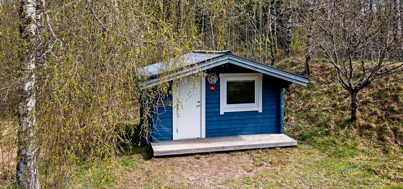 En liten blå trästuga med vit dörr och vitt fönster står på en gräsmatta, omgiven av träd och buskar med tidiga vårlöv.
