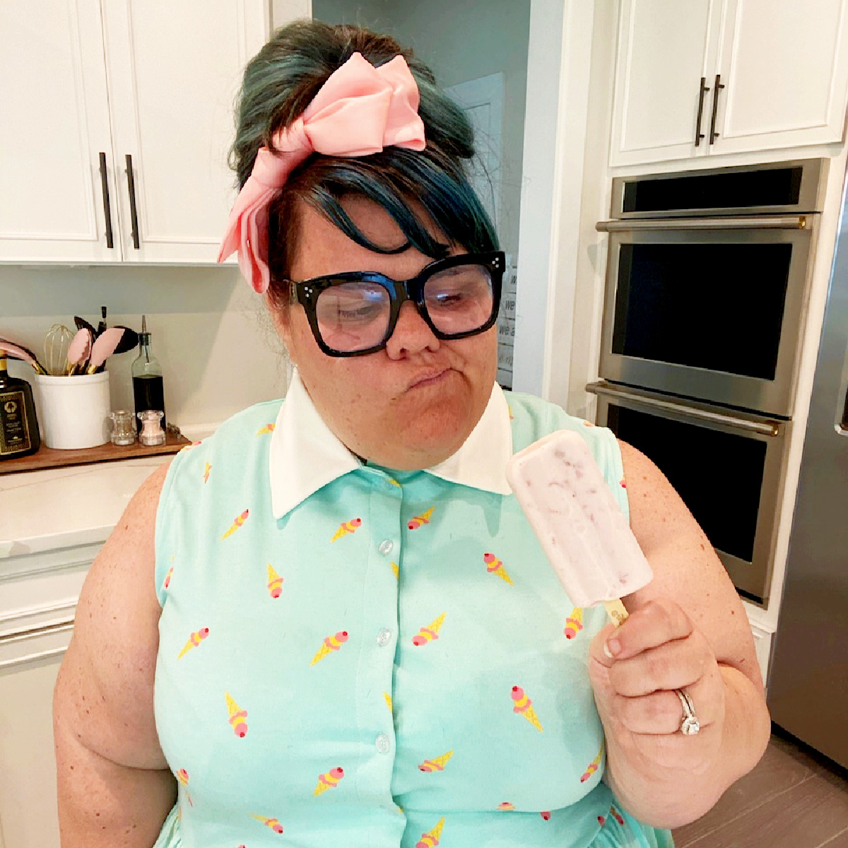 Photo of Liz Davis in her kitchen. She’s wearing an ice-cream-print dress and a pink bow in her hair and is holding a yogurt popsicle.