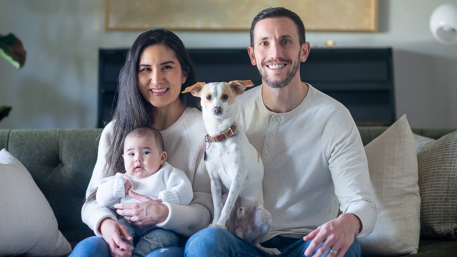 Jocelyn Apodaca Schlossberg is pictured sitting on a sofa with her husband, baby daughter, and their dog.