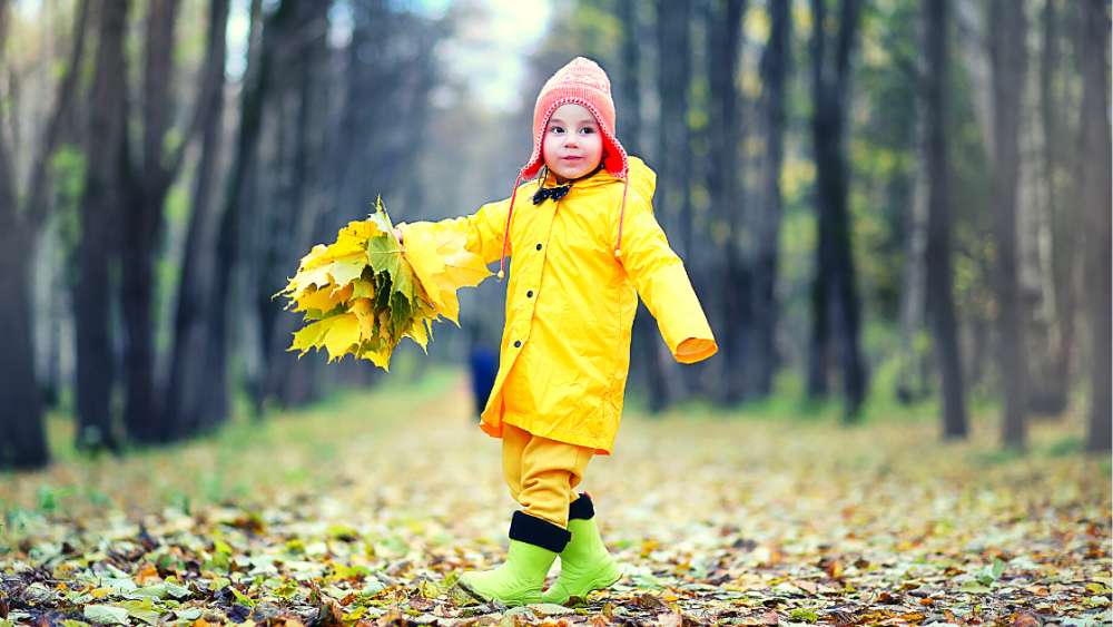 Toddler in yellow raincoat with orange beanie and green boots walking through the forest during Autumn
