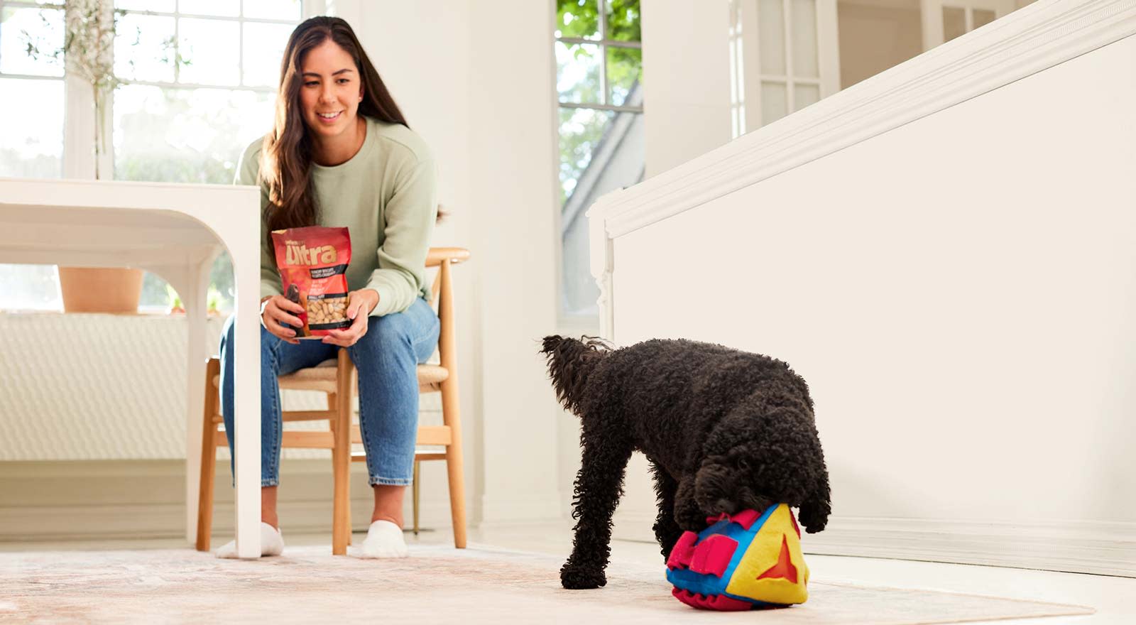 Woman holding a Performatrin ultra treats pack while watching her dog play with a stuffed toy