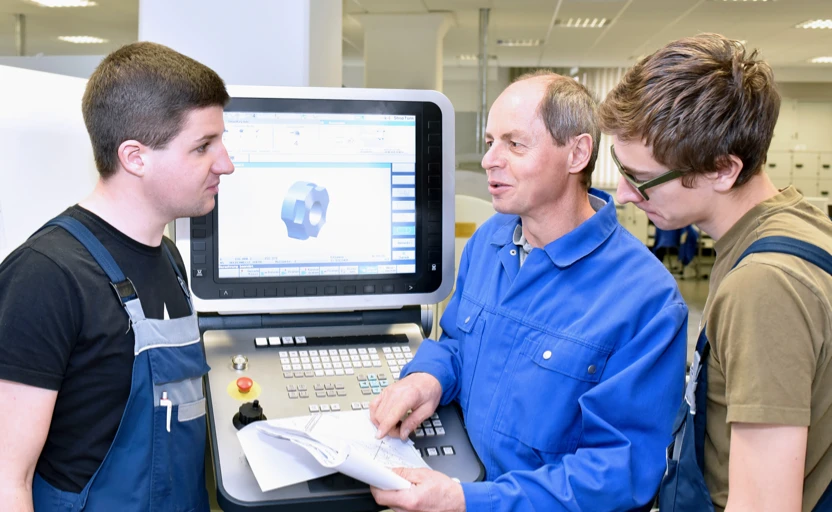 3 people in work overalls standing around a machine, 1 person is explaining how it works to the others