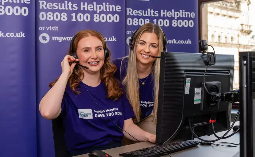 Two women, both wearing purple Results Helpline t-shirts and headsets, smile at the camera.