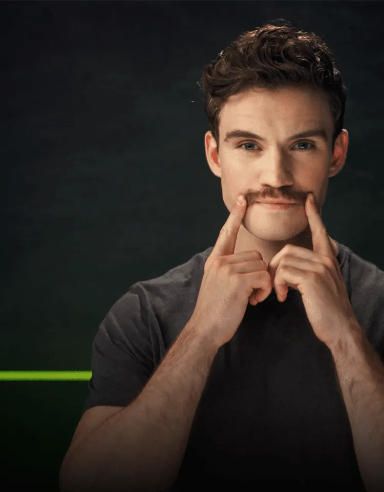 Ein Mann mit Schnurrbart setzt beide Zeigefinger an die Enden seines Schnurrbartes und deutet darauf.