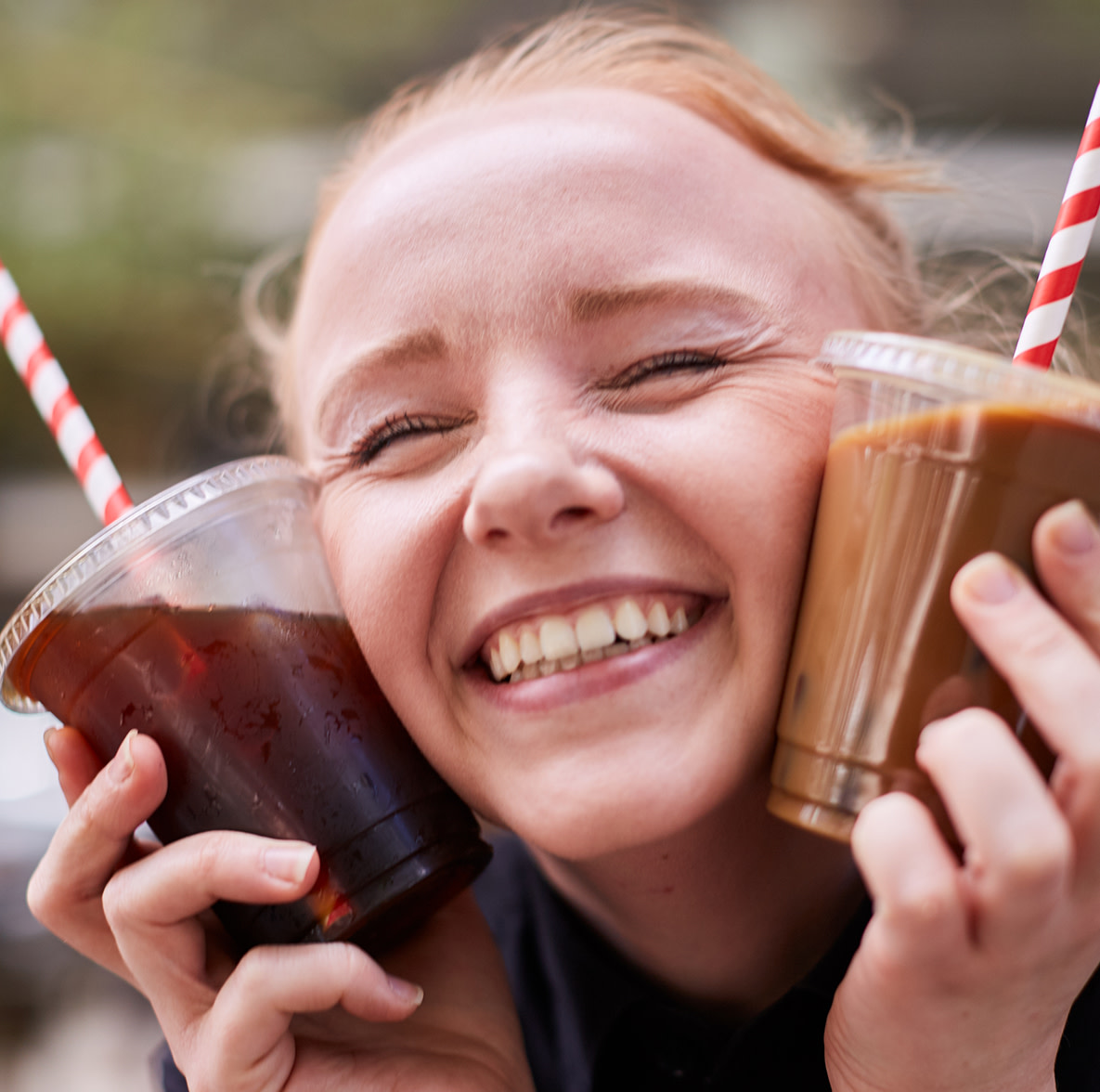 Un jeune femme serrant deux tasses de cafés glacés affiche en grand sourire.