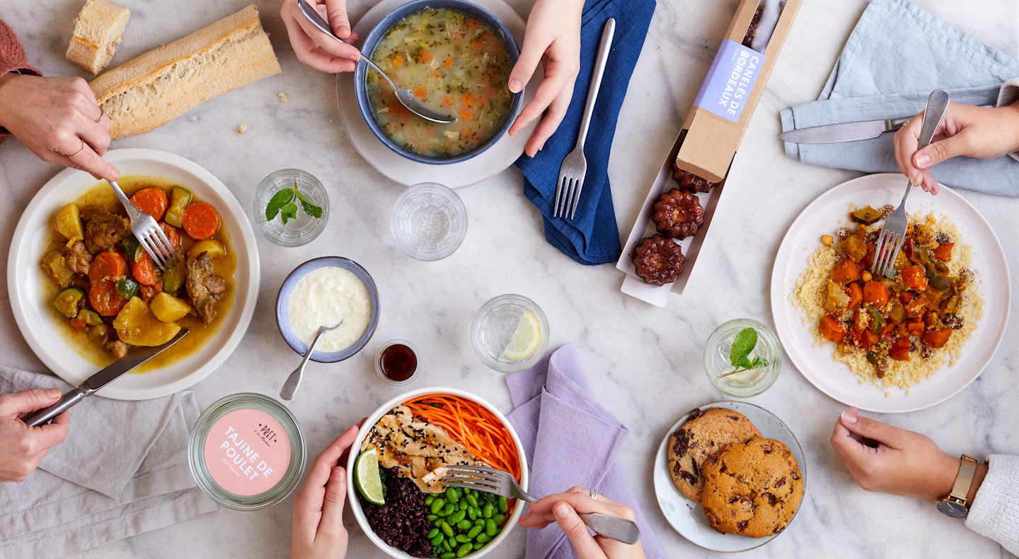 Des personnes dégustent des produits Pret à la Maison : soupes, salade bowl et deux plats chauds sur une table de cuisine avec une baguette de pain, des cookies, des canelés et un bol de riz au lait.
